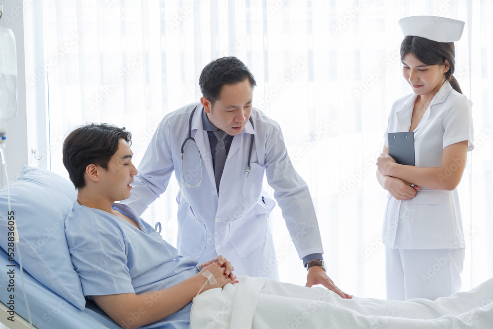 Fototapeta premium Senior doctor and young male patient who lie on the bed while checking symptom, consult and explain with nurse taking note and supporting in hospital wards.