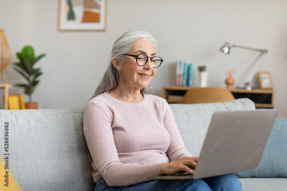 Happy caucasian mature gray-haired female in glasses typing on laptop, watching video in living room interior Happy caucasian mature gray-haired female in glasses typing on laptop, watching video in living room interior