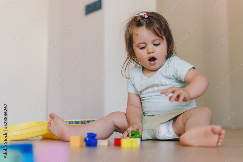 Baby girl playing indoors sitting on floor in playroom at home with constructor. Educational game for baby and toddler. Children build toy house. Creative Baby girl playing indoors sitting on floor in playroom at home with constructor. Educational game for baby and toddler. Children build toy house. Creative