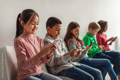 Multiethnic School Kids Using Mobile Phones Sitting Over Gray Background