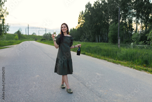 An attractive woman with a bottle and a glass of champagne is walking along the road. Front view.