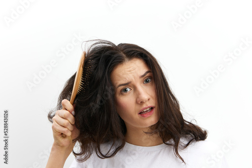 a sad, upset woman is trying to comb her long, dark, tangled hair with a wooden massage comb, standing in a white T-shirt on a white background and making a sad, funny face