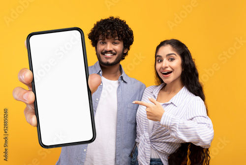 Mobile Offer. Cheerful Indian Couple Pointing At Blank Smartphone With White Screen © Prostock-studio