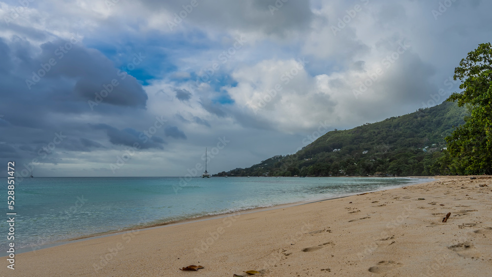 A beach on a tropical island. Footprints in the sand. The turquoise ocean is calm. A green hill against a background of blue sky and clouds. Seychelles. Mahe. Beau Vallon