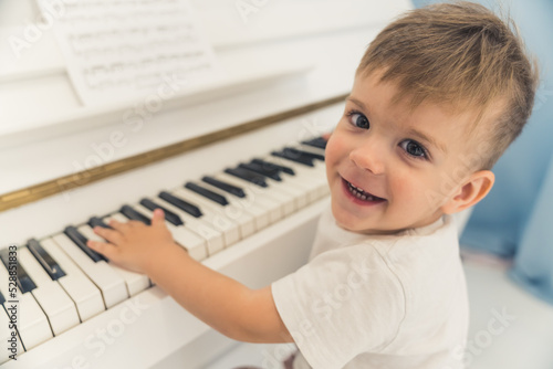 Closeup portrait of an adorable caucasian four-year-old boy playing the piano, looking at camera, and smiling. Casual clothing. Music development. High quality photo