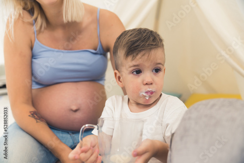 Funny guilty little caucasian baby preschooler boy drinking all of the milk from a cup. Milk marks around the lips. Pregnant mother with visible belly in the background. High quality photo