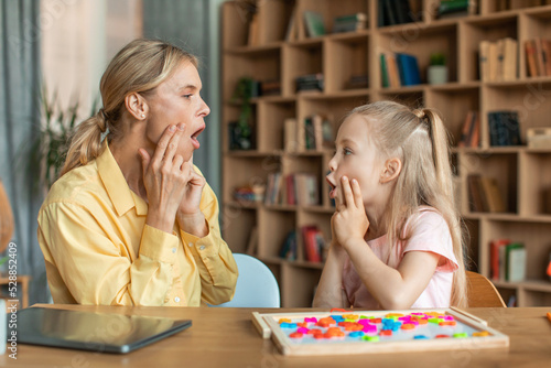 Professional woman speech therapist studying together with pretty little girl, learning practice pronunciation exercises photo