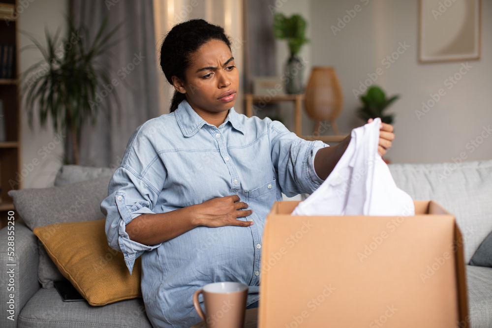 Discontented Pregnant Black Woman Holding Delivered Clothes Unpacking Box Indoor Discontented Pregnant Black Woman Holding Delivered Clothes Unpacking Box Indoor