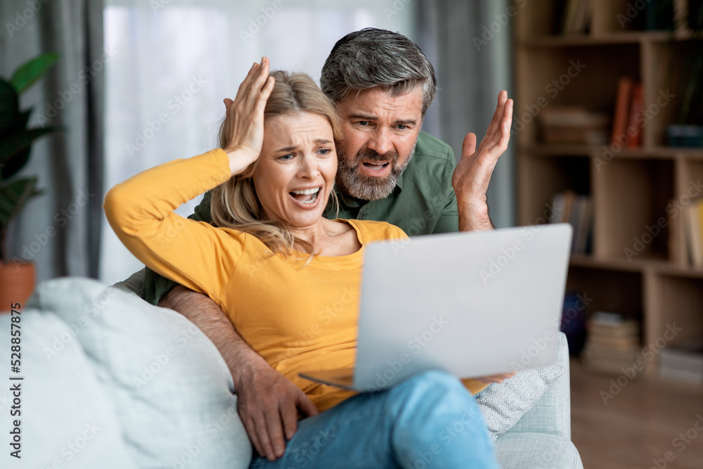Bad News Concept. Shocked Middle Aged Couple Sitting With Laptop On Couch Bad News Concept. Shocked Middle Aged Couple Sitting With Laptop On Couch