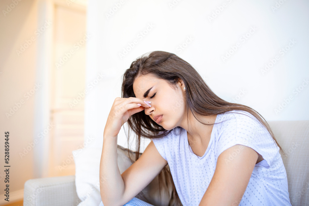 Portrait of an attractive woman sitting on a sofa at home with a headache, feeling pain and with an expression of being unwell. Upset woman sitting on couch feeling strong headache migraine. Portrait of an attractive woman sitting on a sofa at home with a headache, feeling pain and with an expression of being unwell. Upset woman sitting on couch feeling strong headache migraine.