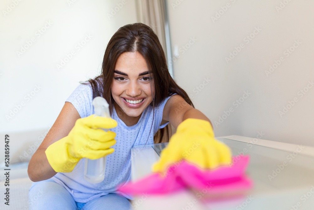 Beautiful young woman makes cleaning the house. Girl rubs dust. Smiling woman wearing rubber protective yellow gloves cleaning with rag and spray bottle detergent. Home, housekeeping concept. Beautiful young woman makes cleaning the house. Girl rubs dust. Smiling woman wearing rubber protective yellow gloves cleaning with rag and spray bottle detergent. Home, housekeeping concept.