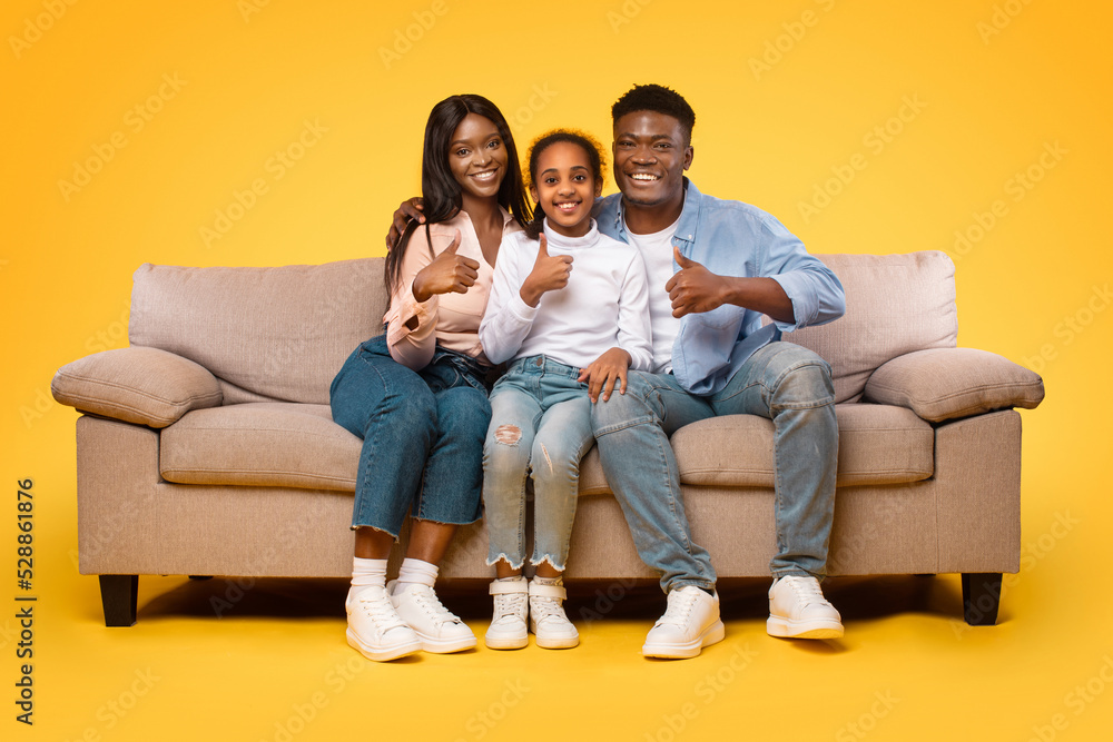 We like it. Happy african american family showing thumbs up gesture, sitting on couch over yellow background, free space