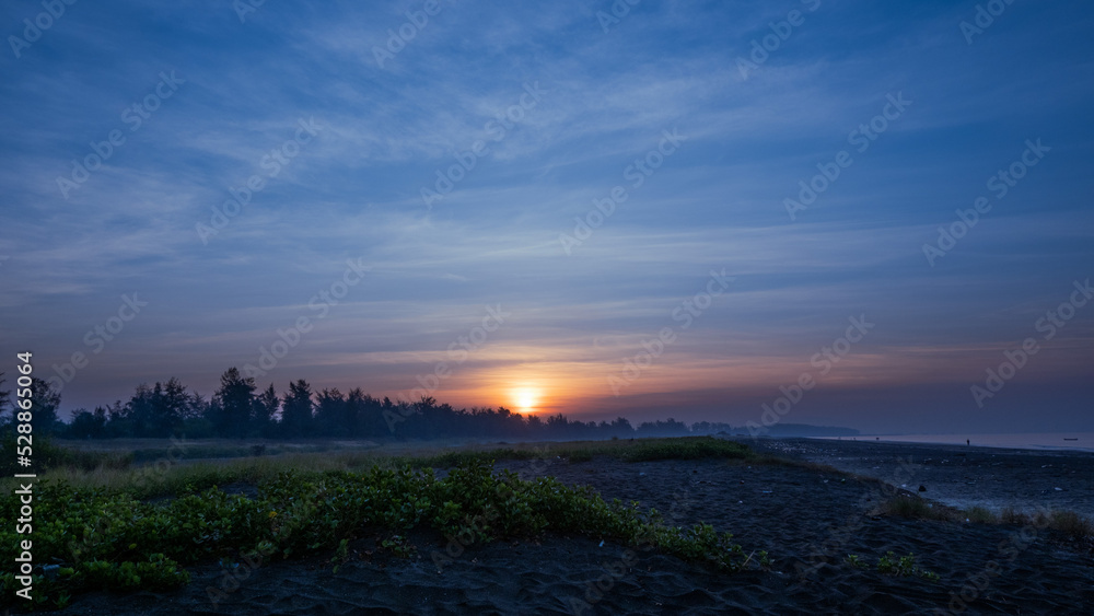 Obraz premium The golden orange sun is rising through the trees behind the sand under the bush at Suruchi beach in Vasai, breaking through the cloud lines of the blue sky.