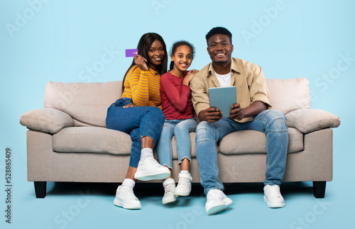 Happy black family of three using digital tablet and credit card for online shopping, sitting on couch, blue background