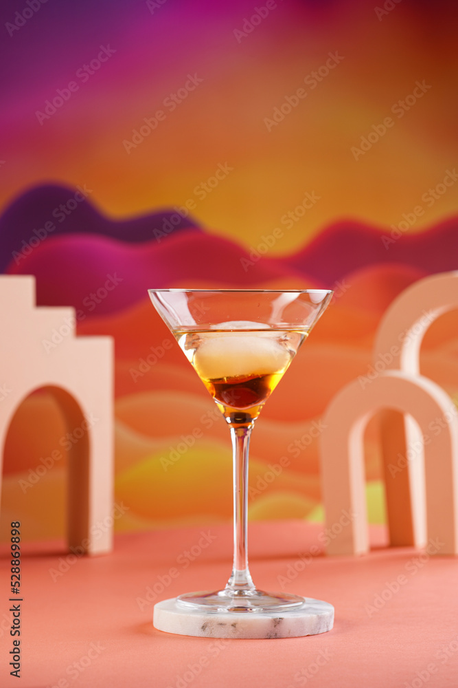 One martini glass with ice cube and brown alcoholic drink on bright colorful red and orange background