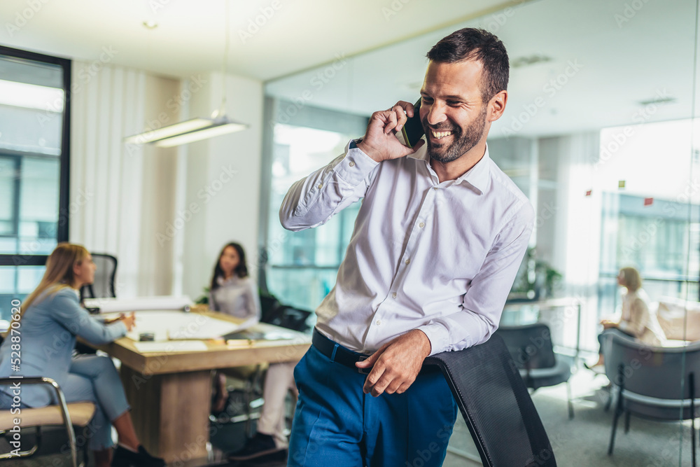 Happy businessman using phone with team discussing project in the background. Happy businessman using phone with team discussing project in the background.