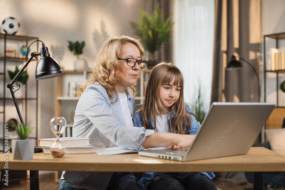 Caucasian young blond woman mother help her beautiful daughter to do homework using laptop computer sitting on table in living room at home. Caucasian young blond woman mother help her beautiful daughter to do homework using laptop computer sitting on table in living room at home.