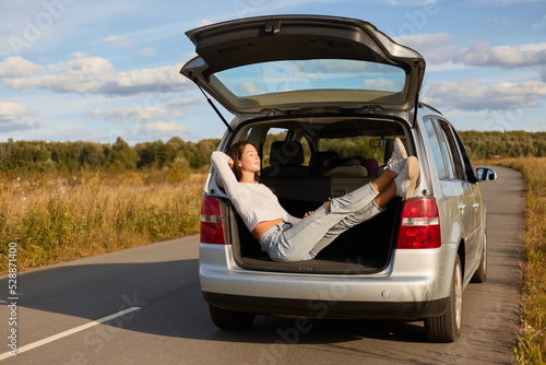 Full length portrait of relaxed woman sitting in the open trunk of a car and basking in the sun, female wearing white shirt and jeans, beautiful female traveling alone. © sementsova321