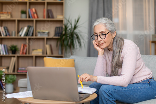 Serious tired busy caucasian senior gray-haired woman teacher in glasses work at laptop in living room interior