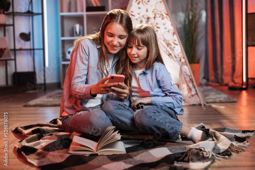 Beautiful cute sisters or friends sitting outside wigwam with smartphone. Happy girls spending evening time together watching favorite movie on phone at home.