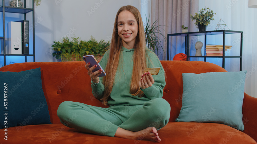 Lovely girl using credit bank card and smartphone while transferring money, purchases online shopping, order food delivery at modern home apartment indoors. Young child kid in living room sit on couch Lovely girl using credit bank card and smartphone while transferring money, purchases online shopping, order food delivery at modern home apartment indoors. Young child kid in living room sit on couch