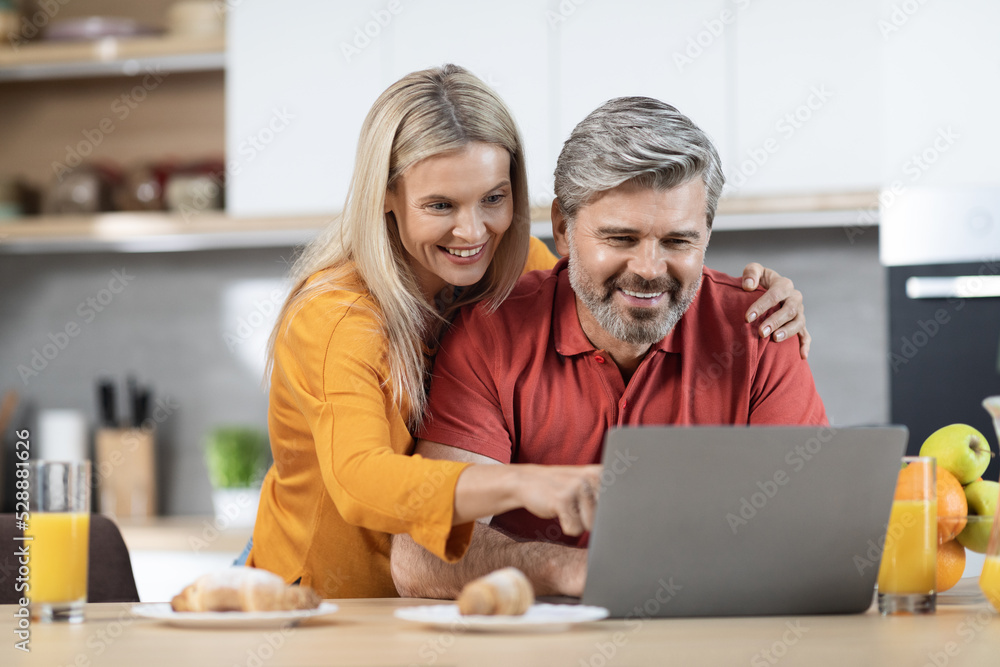 Loving couple enjoying healthy breakfast, using computer, kitchen interior