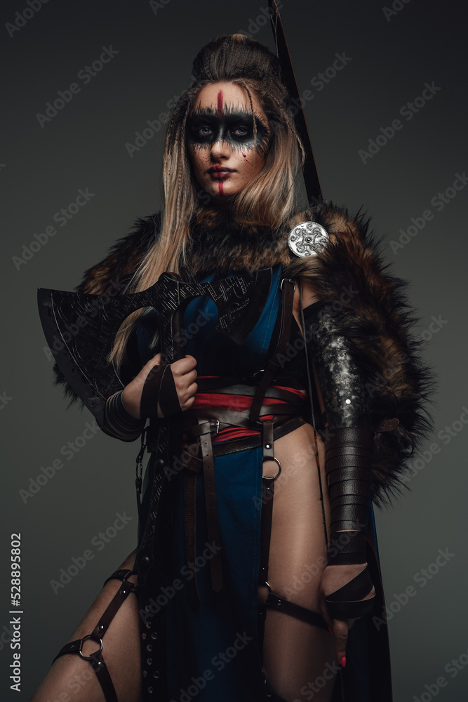 Portrait of female warrior dressed in attire and fur holding axe against grey background.