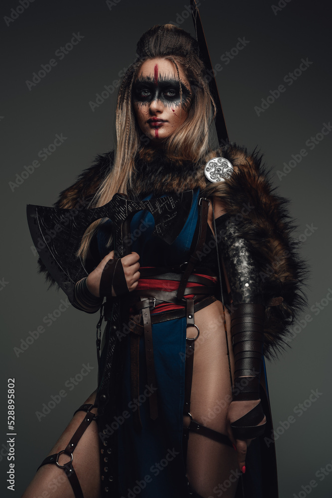 Portrait of female warrior dressed in attire and fur holding axe against grey background. Portrait of female warrior dressed in attire and fur holding axe against grey background.