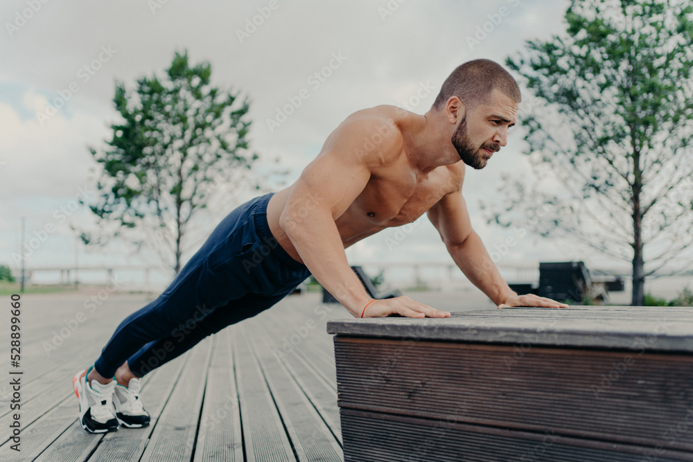Obraz premium Confident muscled sportsman in sport wear doing push up outdoors, works on biceps and trains determination. Strong topless athletic man exercises in open air. People, motivation, training concept