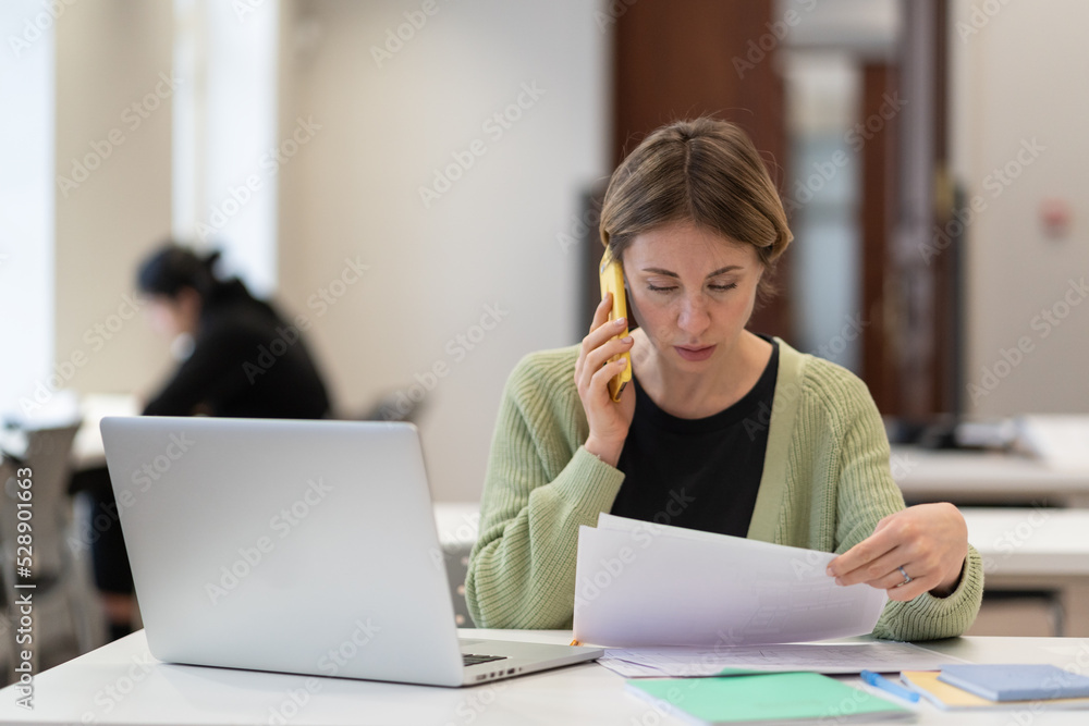 Middle-aged female busy with documents talking on cellphone, working on laptop in campus open space. Focused mature student woman studying preparing for exam in university. Education has no age limit Middle-aged female busy with documents talking on cellphone, working on laptop in campus open space. Focused mature student woman studying preparing for exam in university. Education has no age limit