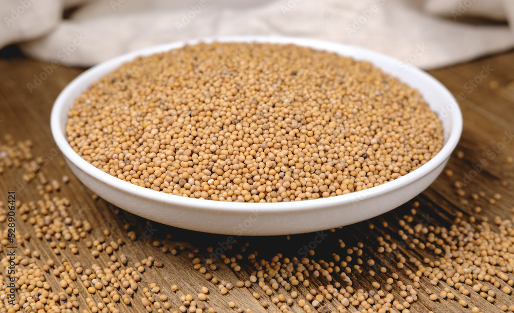 Mustard seeds are poured into a white bowl, standing on a wooden table against a background of light textured and wrinkled fabric from a storage bag. Mustard seeds are poured into a white bowl, standing on a wooden table against a background of light textured and wrinkled fabric from a storage bag.