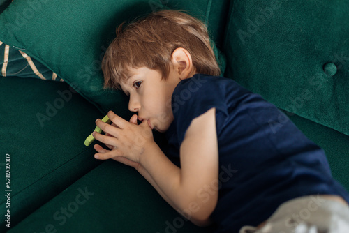 Portrait of serious, scary, sad and depressed little boy lying on green sofa, sucking thumb finger. Children stuttering photo