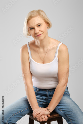 Barefoot senior woman in jeans and tank top sitting on chair in studio