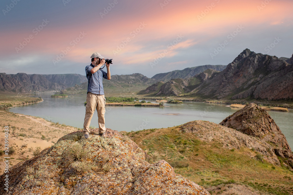 Photographer shoots a landscape from the top of a mountain on the banks of the river on sunset.