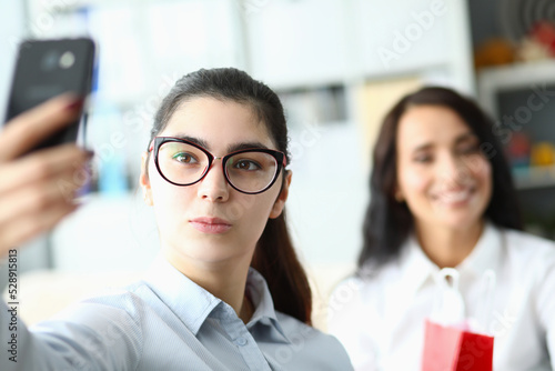 Young woman in glasses holds phone in front of face and smiles at colleagues in office