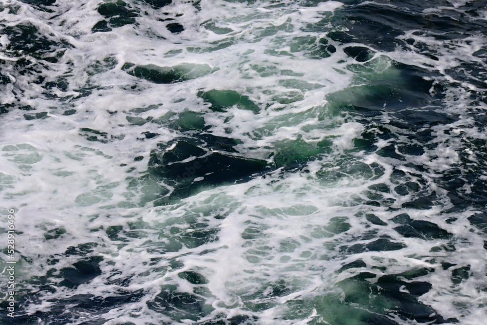 Stern waves with white foam tips on greyish blue sea water, photo taken from aboard ship. Selective focus