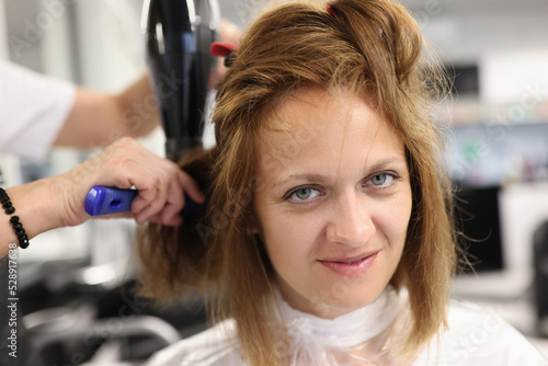 Woman hairdresser dries and straightens client hair with comb and hair dryer in beauty salon