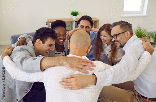 Team of successful positive colleagues laughing and hugging while standing in circle in office. Cheerful business colleagues celebrate their success together. Concept of cohesion and teamwork. photo