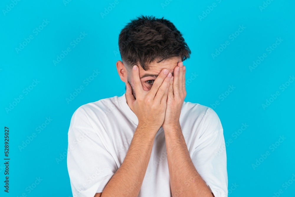 bearded caucasian man wearing white T-shirt over blue background covering face with hands and peering out with one eye between fingers. Scared from something or someone. bearded caucasian man wearing white T-shirt over blue background covering face with hands and peering out with one eye between fingers. Scared from something or someone.