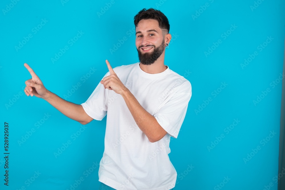 bearded caucasian man wearing white T-shirt over blue background with positive expression, indicates with fore finger at blank copy space for your promotional text or advertisement. bearded caucasian man wearing white T-shirt over blue background with positive expression, indicates with fore finger at blank copy space for your promotional text or advertisement.