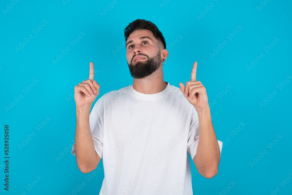 Successful friendly looking bearded caucasian man wearing white T-shirt over blue background exclaiming excitedly, pointing both index fingers up, indicating something. Successful friendly looking bearded caucasian man wearing white T-shirt over blue background exclaiming excitedly, pointing both index fingers up, indicating something.
