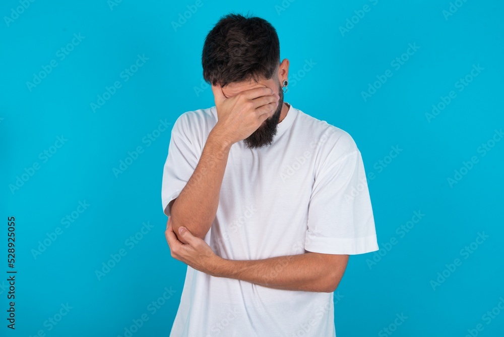 bearded caucasian man wearing white T-shirt over blue background making facepalm gesture while smiling amazed with stupid situation. bearded caucasian man wearing white T-shirt over blue background making facepalm gesture while smiling amazed with stupid situation.