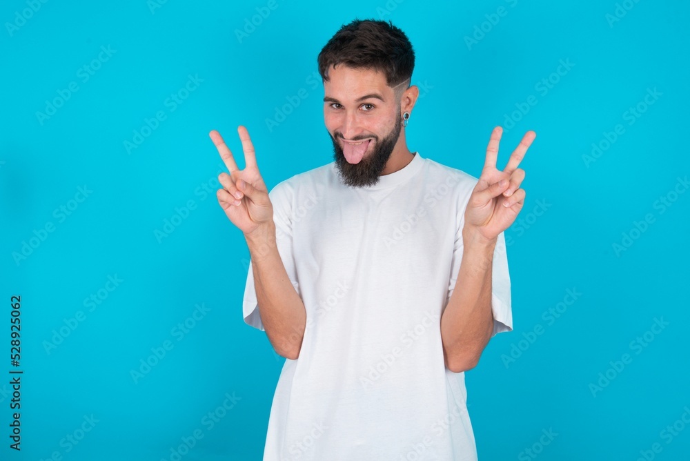 Fototapeta premium bearded caucasian man wearing white T-shirt over blue background with optimistic smile, showing peace or victory gesture with both hands, looking friendly. V sign.