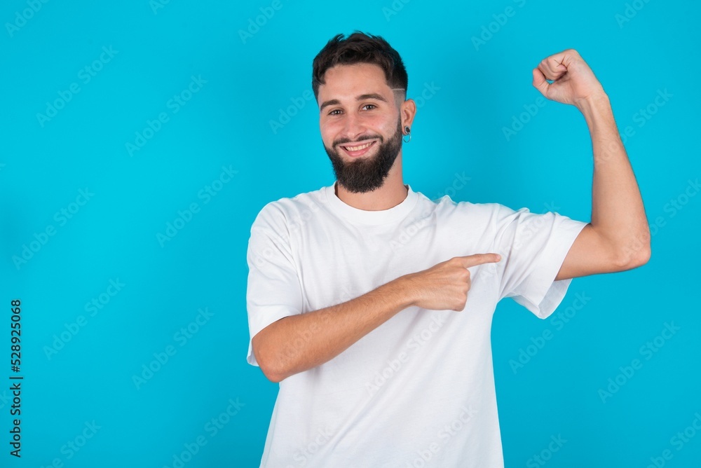 Smiling bearded caucasian man wearing white T-shirt over blue background raises hand to show muscles, feels confident in victory, strong and independent. Smiling bearded caucasian man wearing white T-shirt over blue background raises hand to show muscles, feels confident in victory, strong and independent.