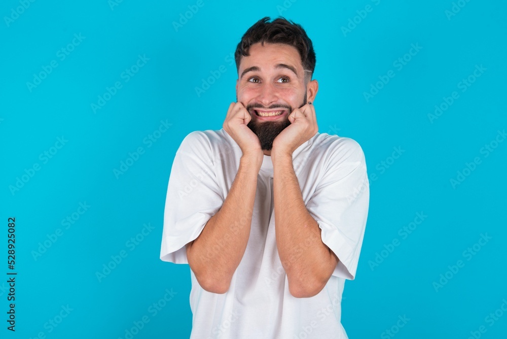 Portrait of bearded caucasian man wearing white T-shirt over blue background being overwhelmed, expressing excitement and happiness with closed eyes and hands near face. Portrait of bearded caucasian man wearing white T-shirt over blue background being overwhelmed, expressing excitement and happiness with closed eyes and hands near face.