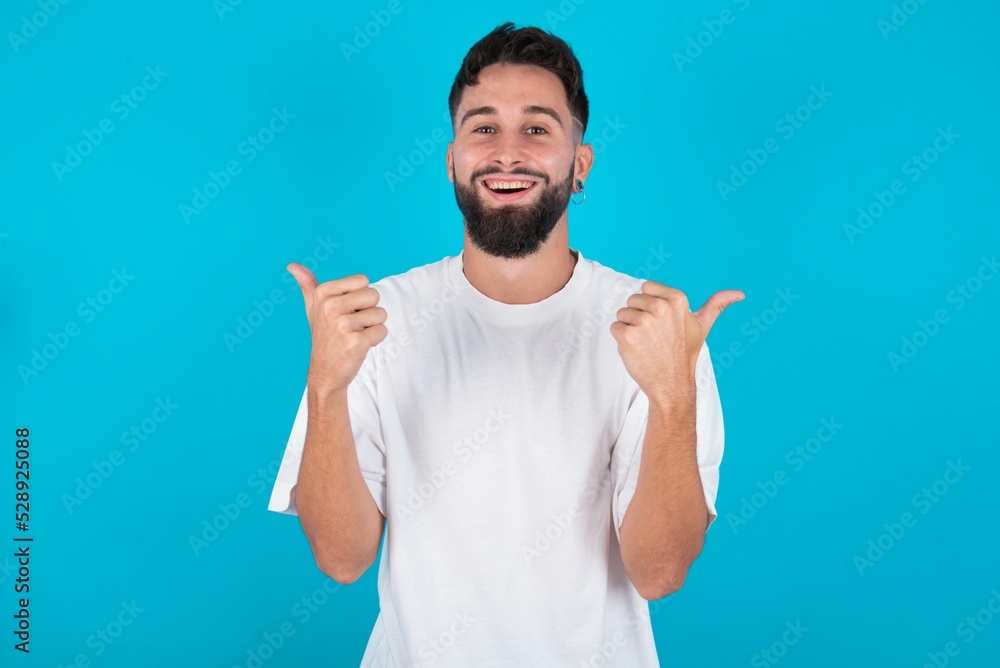 Fototapeta premium Optimistic bearded caucasian man wearing white T-shirt over blue background showing thumbs up with positive emotions. Quality and recommendation concept.