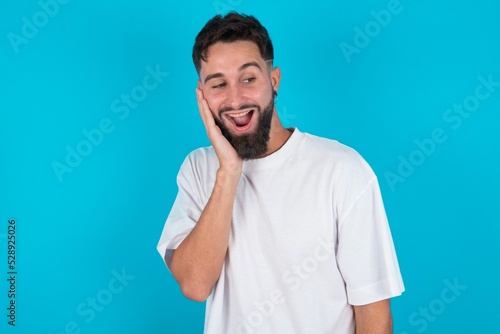 bearded caucasian man wearing white T-shirt over blue background excited looking to the side hand on face. Advertisement and amazement concept.