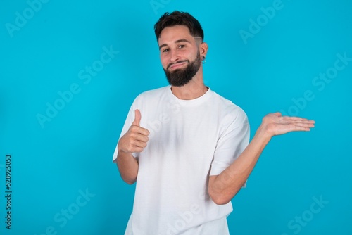 Happy cheerful bearded caucasian man wearing white T-shirt over blue background showing thumb up and pointing with the other hand. I recommend this.