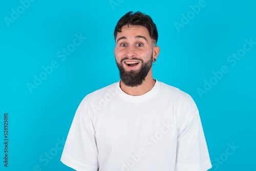 Surprised bearded caucasian man wearing white T-shirt over blue background, shrugs shoulders, looking sideways, being happy and excited. Sudden reactions concept.