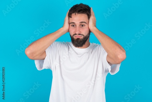 Frustrated bearded caucasian man wearing white T-shirt over blue background plugging ears with hands does not wanting to listen hard rock, noise or loud music. © Jihan